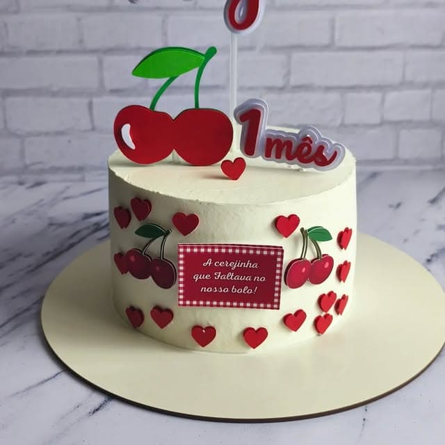 Bolo branco decorado com cerejas de papel e corações vermelhos, com o topo de 1 mês